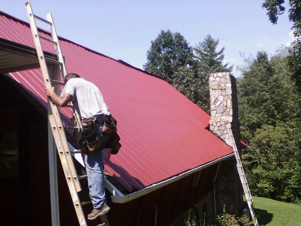 Roofer on a ladder beside a red metal roof and stone chimney — Soffit & Fascia Repair by Roofing Brothers in Milford Square